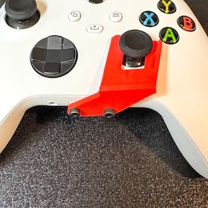 May include: A white Xbox controller with black joysticks and colored buttons. A red accessory is attached to the controller, extending the joystick height. The controller has the Xbox logo and the text "© Kleet3DPrints".