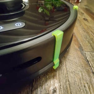 May include: A black robot vacuum cleaner with green bumper guards. The vacuum cleaner is on a wooden surface.