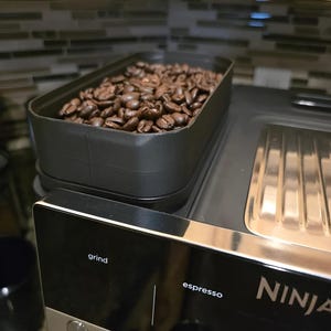 May include: A black espresso machine with a container filled with roasted coffee beans. The machine has a stainless steel trim and the word "grind" is visible. The word "espresso" is also visible.
