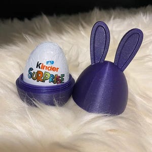 May include: A purple 3D-printed Easter egg holder with bunny ears, next to a Kinder Surprise egg. The egg is white with the words "Kinder Surprise" in colorful letters. The holder is open, revealing the egg.