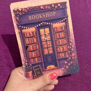 May include: A rectangular card featuring an illustration of a purple bookshop storefront with shelves of books, string lights, pink flowers, a black cat, a sleeping orange cat, and a sign that reads BOOKSHOP and so many books so little time.