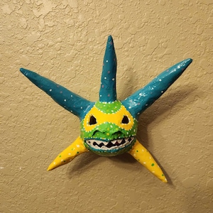 May include: A blue, yellow, and green paper mache mask with a star shape and a toothy grin. The mask has white polka dots on the blue sections and green and yellow polka dots on the yellow sections.