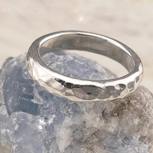 Peut inclure: Une bague de couleur argentée avec une surface martelée et texturée reposant sur une roche cristalline rugueuse bleu clair et blanche. L'anneau métallique présente une finition réfléchissante avec des indentations irrégulières.