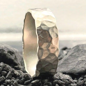 Könnte beinhalten: Ein silberfarbener Metallring mit einer gehämmerten, texturierten Oberfläche. Das Band weist ein unebenes, reflektierendes Finish mit zahlreichen Vertiefungen auf der Außenseite auf und steht aufrecht zwischen dunklen, rauen Gesteinsbrocken.