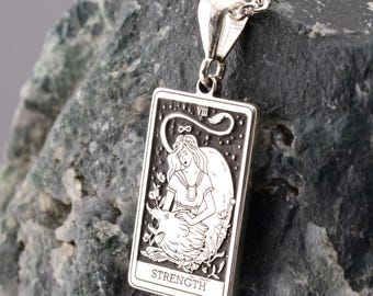 May include: A silver pendant necklace featuring a rectangular charm with the word "STRENGTH" at the bottom. The charm depicts a woman with a lion. The necklace is displayed on a dark green rock.