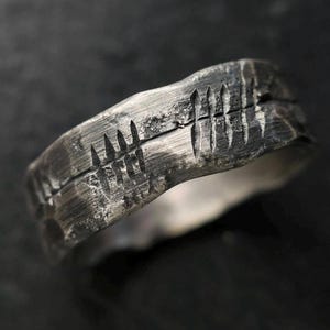 Op de afbeelding: Een donker zilverkleurige ring met een zwaar getextureerd, ruw oppervlak. De band is voorzien van een horizontale lijn die rondom is gegraveerd, onderbroken door verticale streepjes die lijken op een telsysteem in het metaal.