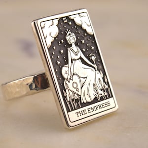 Könnte beinhalten: Silberner Ring mit rechteckiger Oberfläche, die eine Gravur der Tarotkarte Die Kaiserin zeigt. Die Karte zeigt eine Frau in einem Kleid, die ein Zepter hält, mit Sternen und Wolken. Der Ring hat ein gehämmertes Band.