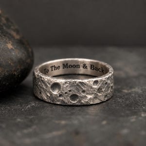 Puede incluir: Un anillo de tono plateado con una superficie texturizada y llena de cráteres que recuerda a la luna. El interior de la banda presenta el texto grabado To The Moon & Back en una fuente oscura y clara, contrastando con el acabado metálico.