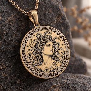 Könnte beinhalten: Eine goldfarbene, runde Anhängerkette mit einer Gravur von Medusa, deren Haar aus Schlangen besteht, umrahmt von einem griechischen Mäander-Muster, einer Mondsichel, Blumen und einem Auge-Symbol, an einer passenden Metallkette.