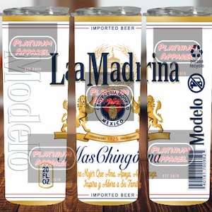 May include: A white can of La Madrina beer with a gold trim and a lion crest. The can features the text "Mas Chingona" and "Imported Beer". The can is 20 fluid ounces.