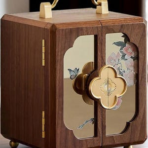 May include: A wooden jewelry box with a handle and brass hardware. The box features two doors with floral and butterfly designs. The box is brown with gold accents and sits on four small gold feet.