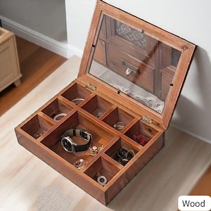 May include: Open wooden jewelry box with a glass top, showcasing various compartments filled with rings, a watch, and other small accessories. The box is made of warm-toned wood and sits on a light-colored surface. The word "Wood" is visible in the bottom right corner.
