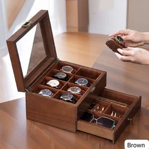 May include: A brown wooden watch box with a glass top, open to reveal multiple compartments holding various wristwatches. A drawer below contains jewelry and sunglasses. The word "Brown" is visible in the bottom right corner.