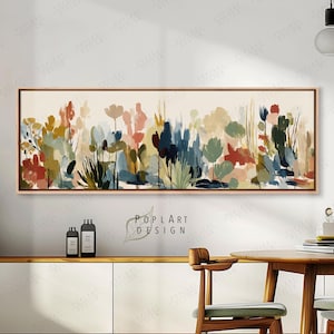May include: Abstract floral painting in a rectangular wooden frame. The artwork features a variety of colorful, stylized flowers and foliage in shades of red, blue, green, and beige. The painting is displayed above a white cabinet and wooden table.