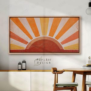 May include: A framed abstract art piece depicting a sunrise with a warm color palette. The artwork features a semi-circle at the bottom with radiating lines in shades of orange, yellow, and white. The frame is a light wood color.