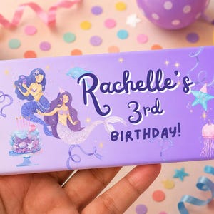 May include: A rectangular chocolate bar wrapper in light purple featuring two mermaids, a birthday cake, a jellyfish, and a star. The text reads Rachelle's 3rd BIRTHDAY! in dark purple script and block lettering with white outlines.