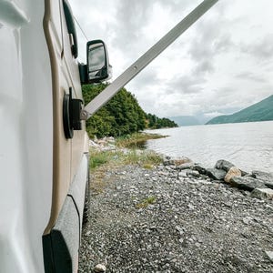 Pode incluir: Uma autocaravana branca estacionada em uma costa de cascalho com vista para um corpo de água e montanhas ao longe. A autocaravana tem um toldo lateral estendido e um espelho lateral.