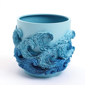 May include: A light blue ceramic pot with a cylindrical shape. The exterior features a detailed wave design in varying shades of blue, creating a textured, three-dimensional effect. The pot is suitable for plants or decorative use.