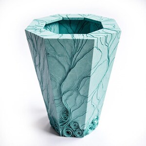 May include: A tall, teal-colored decorative vase with an octagonal top and a flared, geometric shape. The surface features a detailed, raised pattern of swirling lines and organic shapes, resembling tree branches. The base has decorative, rolled paper accents.