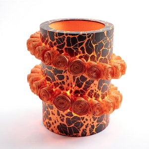 Könnte beinhalten: Zylindrische dekorative Lampe mit einer strukturierten schwarzen und orangefarbenen Außenseite, die an zerbrochene Erde erinnert. Zwei Reihen orangefarbener Papierwirbel umrunden die Lampe. Das Innere leuchtet mit warmem orangefarbenem Licht und erzeugt einen auffälligen visuellen Effekt.