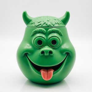 Puede incluir: Un tarro de galletas de cerámica verde con forma de cara de monstruo de dibujos animados. El tarro tiene ojos negros grandes, una boca sonriente con una lengua rosa y cuernos pequeños. La tapa tiene una textura que simula pelo.