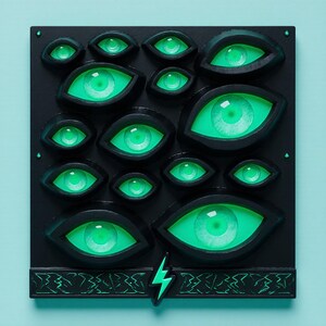 May include: A black square art piece featuring multiple glowing green eyes of varying sizes. The eyes are set against a black background, with a decorative border and a lightning bolt accent at the bottom. The piece is mounted on a light blue background.