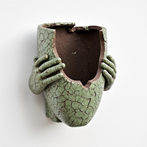 May include: A green ceramic wall planter with a cracked, textured surface. The planter is shaped like a broken vessel, with two hands appearing to hold it. The interior is a contrasting brown color.