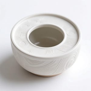 May include: A white ceramic bowl with a circular design and a central opening. The bowl features a textured pattern of curved lines and circles, and a natural-colored base. The design is minimalist and modern.