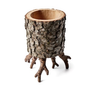 Może przedstawiać: Cylindryczna drewniana donica z naturalną korą na zewnątrz i pustym wnętrzem. Donica jest podparta przez kilka nóżek przypominających korzenie, tworząc unikalny, organiczny design. Idealna do małych roślin.