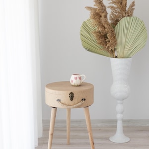 Puede incluir: Una mesa auxiliar redonda, de color marrón claro, con tres patas de madera. La mesa tiene un diseño tipo maleta con asa y cierre. Encima hay una taza blanca pequeña con diseños de tulipanes rosas. Un jarrón alto blanco con plantas secas en el fondo.