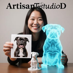 May include: The image features a smiling person holding a framed picture of a black and white dog. A white dog figurine and a glowing blue dog sculpture are also present. The text "ArtisanTeestudioD" is at the top.