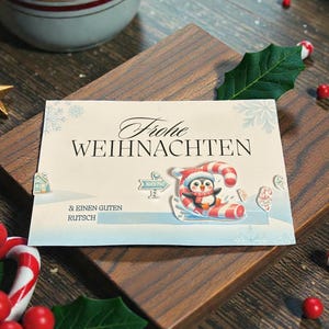 May include: A holiday greeting card with the words "Frohe Weihnachten" and a penguin on a candy cane sled. The card is on a wooden surface, surrounded by candy canes, holly, and a cup of hot chocolate.
