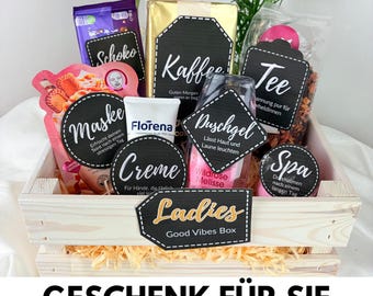 Op de afbeelding: Een houten cadeaukistje gevuld met diverse zelfzorgartikelen, waaronder een gezichtsmasker, koffie, thee en lotion. De doos heeft een "Ladies Good Vibes Box" label en de tekst "GESCHENK FÜR SIE" onderaan.