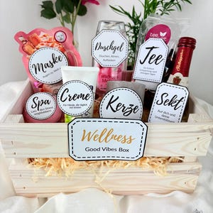 Könnte beinhalten: Eine Holzkiste gefüllt mit Spa-Produkten, darunter eine Gesichtsmaske, Creme, Badebombe, Kerze, Tee und Sekt. Die Kiste trägt das Schild "Wellness Good Vibes Box". Rosa Blumen und Grünzeug sind im Hintergrund zu sehen.