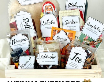 Op de afbeelding: Een houten kist gevuld met kerstartikelen, waaronder een fles Glühwein, een kruik, chocolade, sokken en peperkoek. De kist heeft verschillende cadeaulabels met Duitse tekst. De woorden "WEIHNACHTSKORB" en "12 Etiketten mit Bibelversen" zijn afgedrukt.