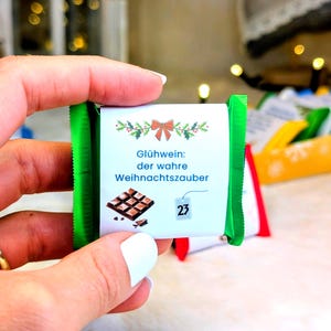 Op de afbeelding: Een kleine chocoladereep met groene verpakking wordt in een hand gehouden. Het etiket toont een strik, hulst en tekst in het Duits. De reep is gemarkeerd met nummer 23, wat wijst op een adventskalender-thema.