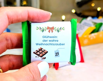 Op de afbeelding: Een kleine chocoladereep met groene verpakking wordt in een hand gehouden. Het etiket toont een strik, hulst en tekst in het Duits. De reep is gemarkeerd met nummer 23, wat wijst op een adventskalender-thema.