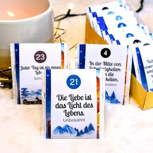 Könnte beinhalten: Nahaufnahme von kleinen, weißen Päckchen mit blauen und goldenen Akzenten, die jeweils eine Nummer und deutschen Text zeigen. Die Päckchen sind in der Nähe einer brennenden Kerze und einer Schachtel mit ähnlichen Päckchen angeordnet.