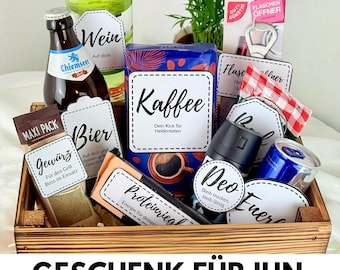 Op de afbeelding: Een houten cadeaupakket gevuld met diverse items, waaronder een fles wijn, bier, koffie, kruiden en een flesopener. De mand heeft labels met Duitse tekst. De tekst "GESCHENK FÜR IHN" wordt onderaan weergegeven.
