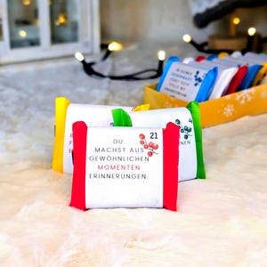 Può includere: Un calendario dell'Avvento con regali confezionati. I regali sono bianchi con bordi colorati e presentano testo e illustrazioni. Il testo su un regalo recita "DU MACHST AUS GEWÖHNLICHEN MOMENTEN ERINNERUNGEN."