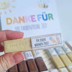 Puede incluir: Una mano sosteniendo una barra de chocolate de color claro con la palabra "merci" grabada. La barra está parcialmente envuelta con una etiqueta que dice "FÜR JEDEN WARMEN TROST" y presenta ilustraciones de lápices. En el fondo, una tarjeta dice "DANKE FÜR" en letras coloridas.