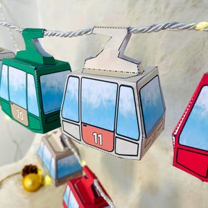 Könnte beinhalten: Eine Girlande aus bunten Papier-Seilbahnkabinen, jede mit einer einzigartigen Nummer. Die Kabinen sind in Grün, Rot und Beige gehalten, mit blauen Himmelsgrafiken in den Fenstern. Die Girlande ist an einem silbernen Seil befestigt, für Dekorationen.