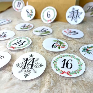 Puede incluir: Números blancos y ovalados para calendario de adviento con numerales negros e ilustraciones en acuarela. Algunos números están rodeados de coronas verdes con lazos rojos, mientras que otros presentan diseños botánicos. Los números están dispersos sobre una superficie suave y clara.