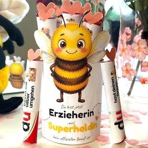 Op de afbeelding: Een cilindrische geschenkhouder met een cartoonbij-illustratie en roze papieren hartjes. De tekst luidt: Du bist jetzt Erzieherin, weil Superheldin kein offizieller Beruf ist. Twee chocoladerepen zijn bevestigd met Duitse tekst erop.