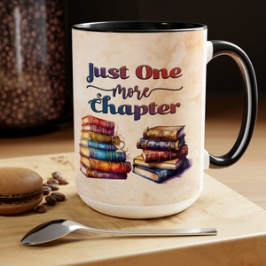 Könnte beinhalten: Weißer Keramikbecher mit schwarzem Innenraum und Henkel. Der Becher zeigt den Text "Just One More Chapter" mit Illustrationen von gestapelten Büchern in verschiedenen Farben. Ein Löffel und ein Gebäck befinden sich im Vordergrund.