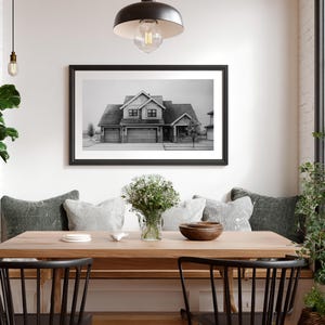 Może przedstawiać: Jadalnia z drewnianym stołem i czarnymi krzesłami. Czarno-biała fotografia domu w ramie wisi na ścianie. St&oacute;ł jest ozdobiony poduszkami, wazonem z kwiatami i drewnianą miską, tworząc przytulną atmosferę.