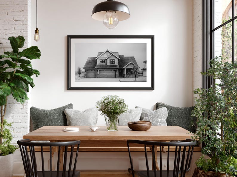 Może przedstawiać: Jadalnia z drewnianym stołem i czarnymi krzesłami. Czarno-biała fotografia domu w ramie wisi na ścianie. St&oacute;ł jest ozdobiony poduszkami, wazonem z kwiatami i drewnianą miską, tworząc przytulną atmosferę.