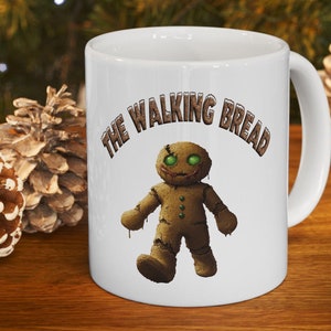 Puede incluir: Taza de cerámica blanca con un hombre de jengibre marrón con ojos verdes y una sonrisa espeluznante. La taza tiene el texto "The Walking Bread" impreso.
