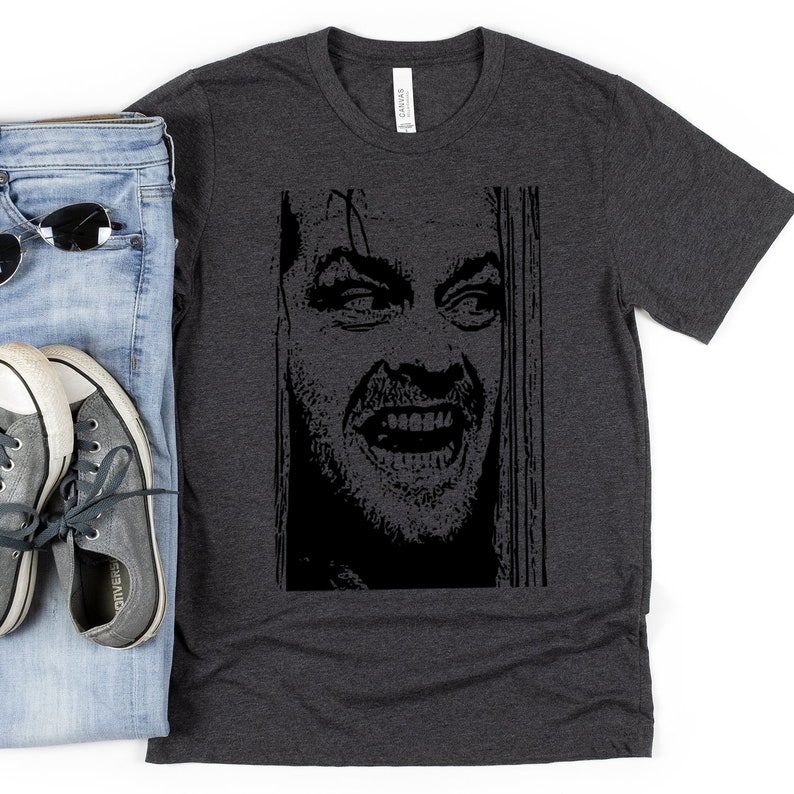 以下が含まれることがあります： ダークヘザーグレーのTシャツに、映画「シャイニング」のジャック・ニコルソン演じるキャラクターの白黒グラフィックプリントが施されています。