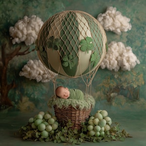 Könnte beinhalten: Ein neugeborenes Baby, eingewickelt in grünen Stoff, liegt in einem Weidenkorb unter einem grün-cremefarbenen Heißluftballon, der mit Kleeblättern verziert ist. Die Szene spielt sich vor dem Hintergrund von Wolken und einem gemalten Wald ab.